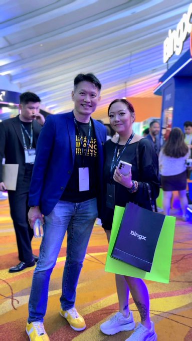 Two people pose together at what appears to be a tech conference; the woman holds a shopping bag with the text 'BingX' visible.