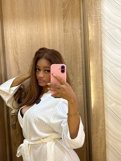A woman takes a mirror selfie in a white waffle-textured robe, holding a pink phone in a wood-paneled room. The image appears to be a casual "GM" (good morning) post featuring a lifestyle shot paired with the $RIVER ticker and related project tags.