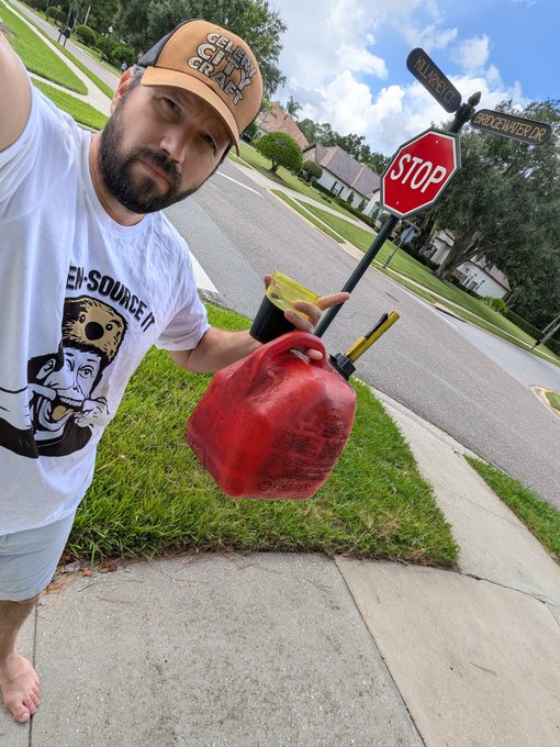 A man in a white 'Open Source' t-shirt and a 'Get Your Crunch On' cap stands barefoot on a sidewalk holding a red gasoline can, gesturing toward a fallen stop sign at the intersection of Allbrook Dr and Creekwater Dr.