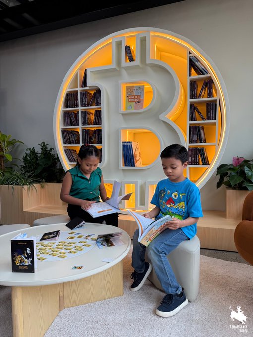 Two children sit at a curved white table reading books in a room featuring a large circular bookshelf shaped like a Bitcoin '₿' symbol with orange backlighting.
