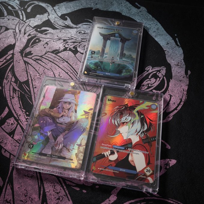 Three Azuki TCG trading cards in protective cases are displayed on a black and white patterned mat, featuring "Hydromancy Gate," "Shao," and another holographic character card (with "70km" visible), along with what appears to be artist signature "IKZ" at the bottom, tying into the post's mention of the Azuki trading card game and the tag "IKZ."