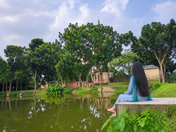 A woman with long dark hair sits on a stone ledge by a pond, looking out at a lush green landscape with large trees and traditional buildings under a partly cloudy sky.