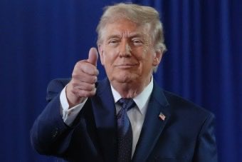 Donald Trump in a dark suit and blue tie giving a thumbs-up against a blue curtain backdrop.
