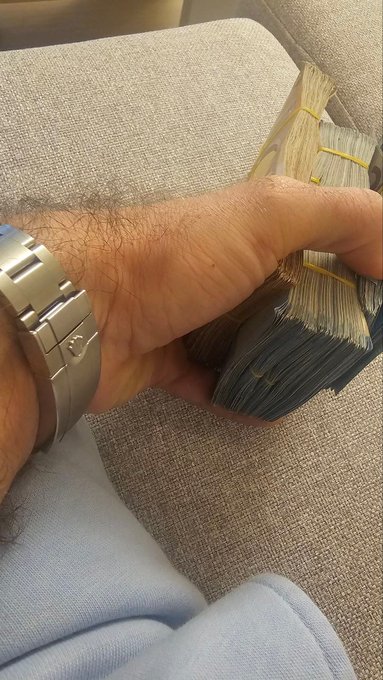 A close-up photo shows a man's hairy arm and hand clutching a thick stack of banded US dollar bills while wearing a metal link bracelet.