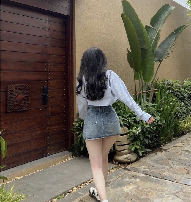 A photograph of a woman with long wavy black hair, seen from behind, wearing a white long-sleeve blouse and a denim mini skirt as she walks along a stone path beside a wooden door and tropical plants.