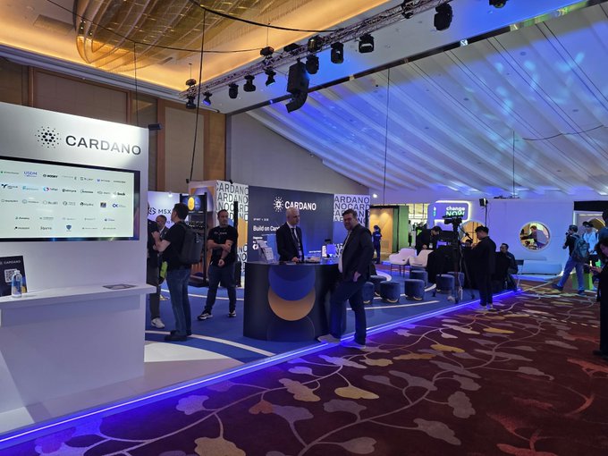 A Cardano-branded exhibition booth at a crypto conference, with displays, attendees, and stage lighting visible.