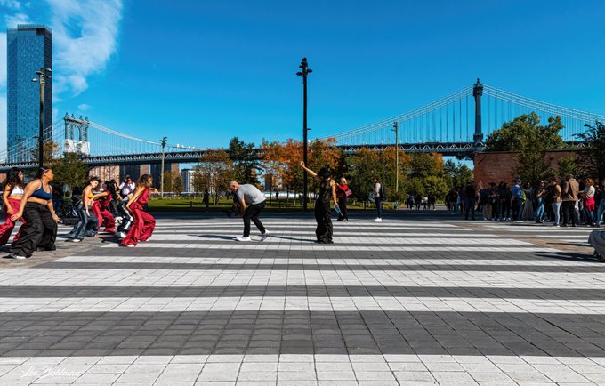 Dancers perform outdoors in a plaza under a clear blue sky, with a large suspension bridge and a crowd in the background.