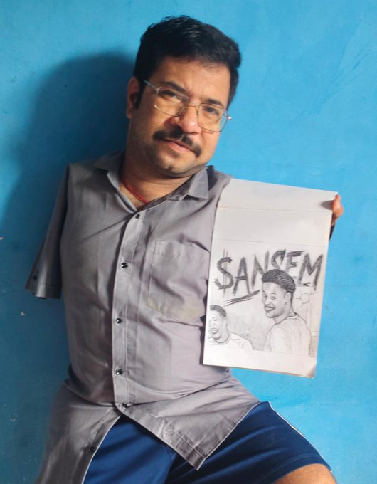 A man named Dhiraj poses against a blue wall, proudly displaying his pencil-drawn artwork of "$ANSEM" featuring two portraits. The sketch relates to his post about creating leg art to support the $ANSEM coin, showcasing his dedication as an artist in the crypto community.