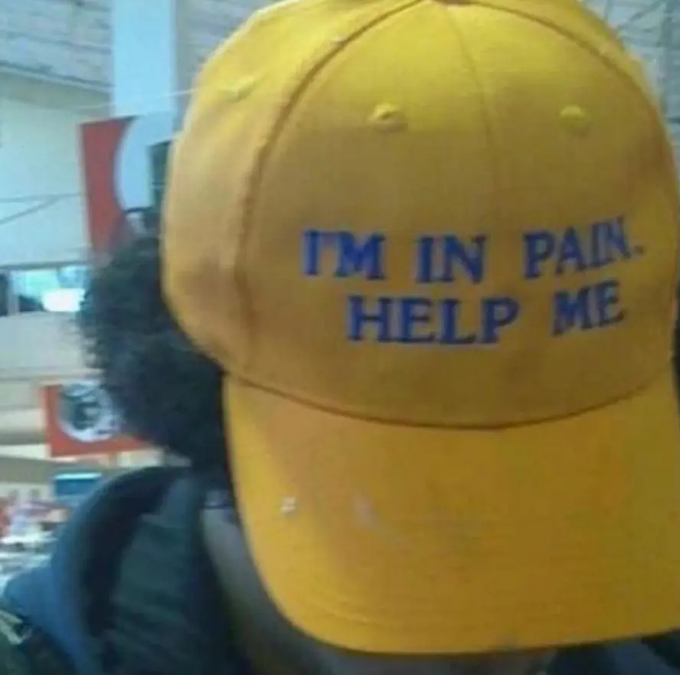 A person wearing a yellow cap with the embroidered text 'ITM IN PAIN. HELP ME' in blue letters.