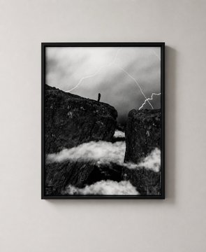 A black-framed black and white photograph hanging on a wall, depicting a dramatic stormy landscape with lightning over a cliff and a tiny figure standing on the edge.