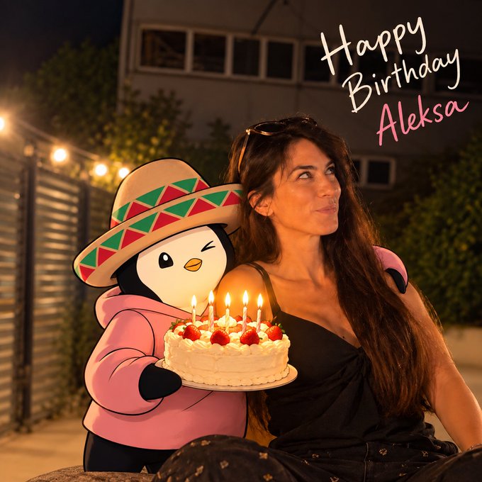 A woman with long dark hair wearing a black dress poses beside a cutout of a penguin wearing a sombrero, with 'Happy Birthday Aleksa' written in the top right corner and a strawberry birthday cake with lit candles in the foreground.