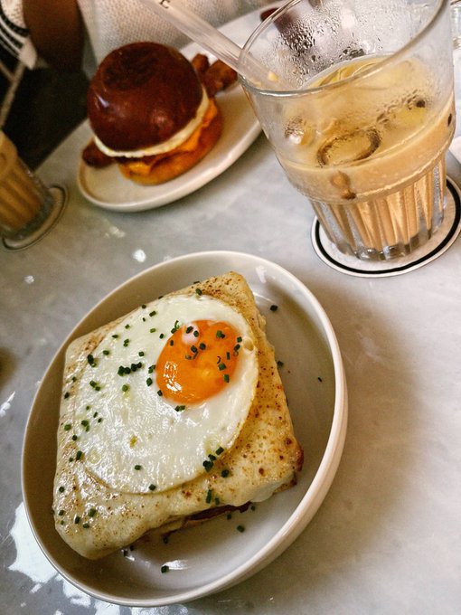 A food photograph showing a croissant sandwich topped with a fried egg and chives in a white bowl, with a glass of water and a small burger visible in the background.