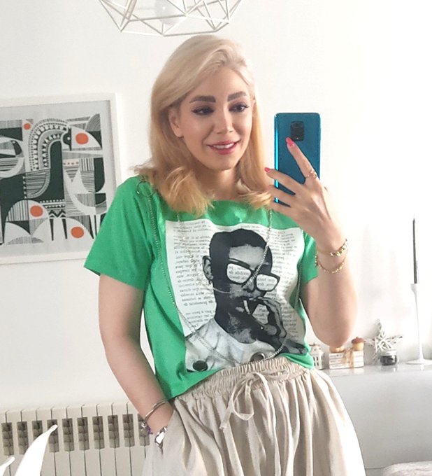 A blonde woman takes a mirror selfie with a blue phone, wearing a green t-shirt featuring a black-and-white printed portrait, paired with a beige elastic-waist skirt, in a bright room with abstract art and a chandelier.