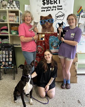 Three people posing with two dogs inside a pet store or shelter in front of a 'Change dog lives one paw at a time!' banner featuring a dog wearing sunglasses with 'WOOF' speech bubble.