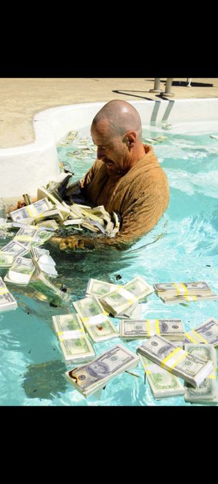 A bald man in an orange robe sits in a pool filled with floating bundles of US dollar bills.