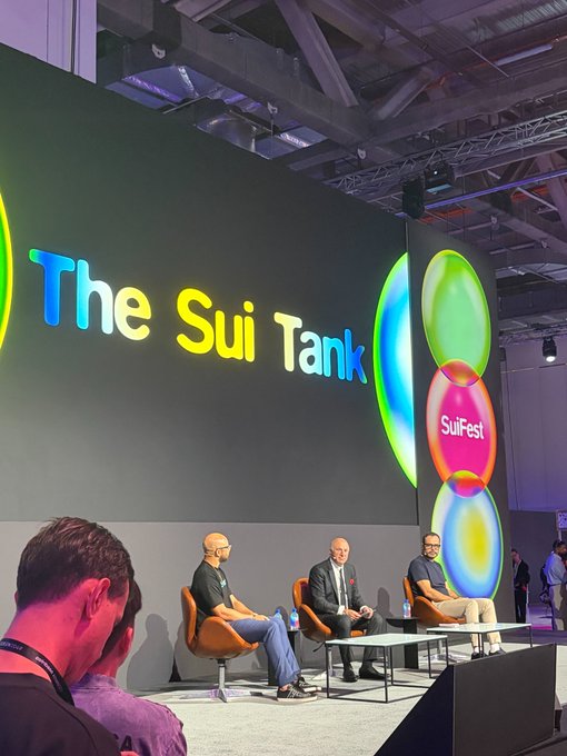 A panel discussion at "The Sui Tank" event, with the "SuiFest" logo displayed on a large screen, showing three men seated on stage in what appears to be a Web3 conference setting. The image captures the lively on-stage atmosphere of Sui's Shark Tank-style pitch event from SuiFest, where industry figures like Kevin O'Leary ("Mr. Wonderful") were brought in as judges.