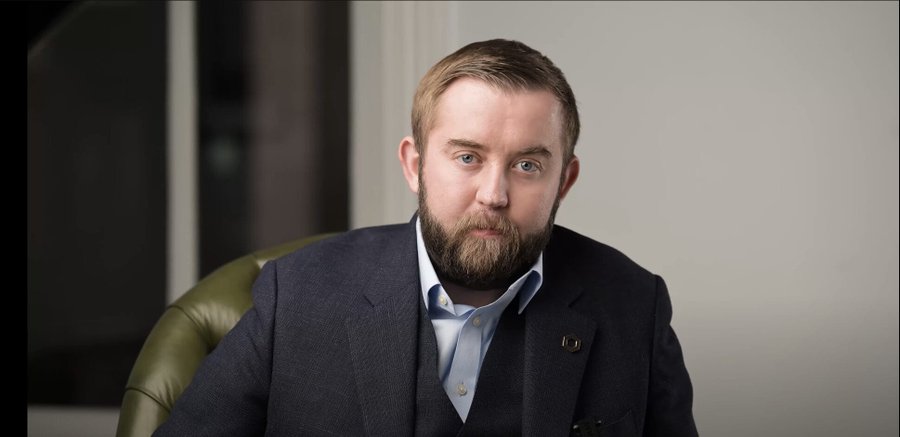 A bearded man with light brown hair, wearing a dark blazer over a light blue collared shirt, sits in a green leather chair against a neutral background, photographed in a professional headshot style. The image appears to be a portrait of a company executive, likely featured in connection with Chainlink's announcement of the CCIP 2.0 launch.