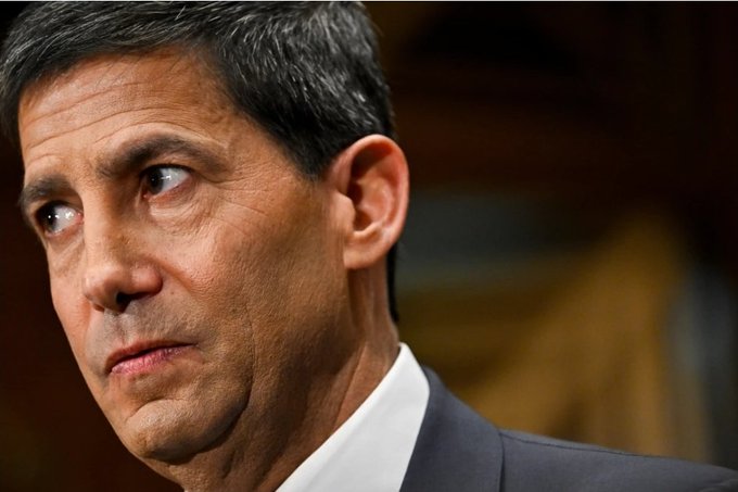 A close-up portrait of a serious-looking man with short gray hair, wearing a dark suit and white shirt, gazing off to the side.
