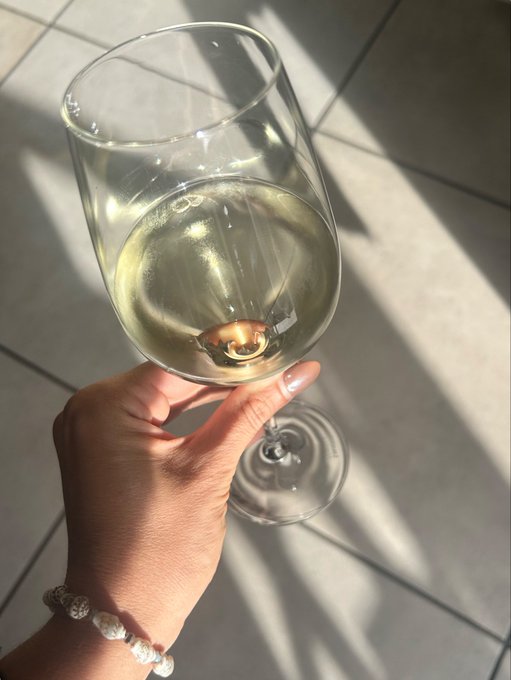 A hand with a beaded bracelet holds a glass of white wine in sunlight, casting shadows on a tiled floor.