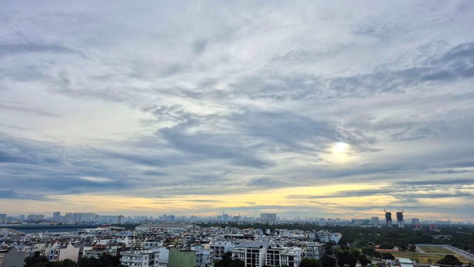 A panoramic cityscape at dusk captures a sprawling urban skyline beneath an expansive cloudy sky, with the sun glowing softly through the clouds — matching the post's early-morning wake-up vibe and anticipation of Elon Musk's email.