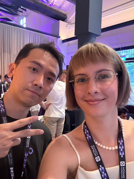 A man and woman take a selfie at the Monad Open event in Singapore during Token2049 week, with lanyards displaying "MONAD" and "TOKEN2049" visible around their necks. The casual pose and event badges reflect the networking atmosphere of the crypto conference.