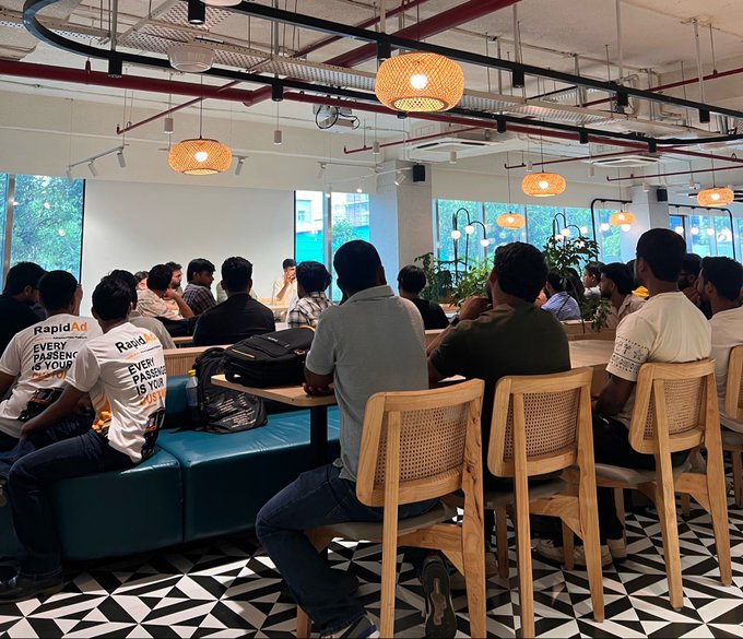 A crowded indoor gathering at a co-working or event space where attendees wearing 'RapidAd' shirts are seated, listening to a speaker at the front.