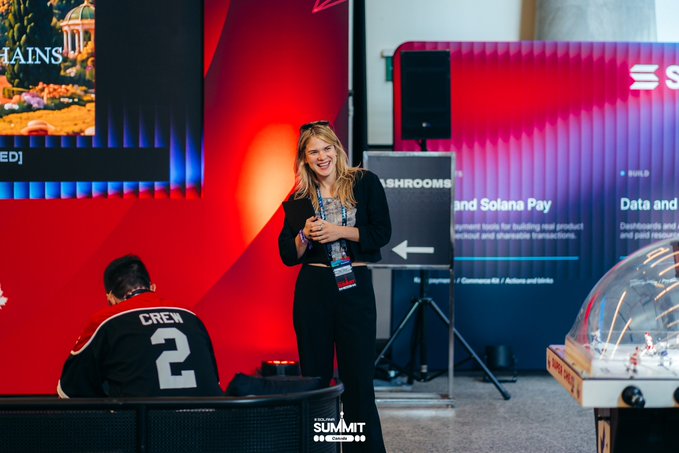 A blonde woman stands at a 'SUMMIT' conference, smiling while holding a drink, next to a banner about Solana Pay and a person wearing a 'CREW' jersey with the number 2.
