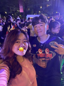 A nighttime selfie of a smiling young woman with a 'crown' sticker and a young man in a black 'V' jersey, posing among a crowd.