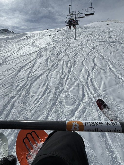A skier captures a first-person view riding a chairlift over a tracked-out snowy slope, with a Bitcoin sticker on the safety bar reading "make war unaffordable." The scene ties into the post's hopeful message from a Chilean ski resort, using the literal chairlift moment as a backdrop for reflecting on global uncertainty and wishing for peace.