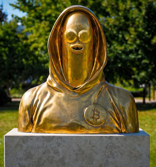 A gold statue of a hooded figure wearing sunglasses with a Bitcoin symbol on its chest, displayed on a pedestal.