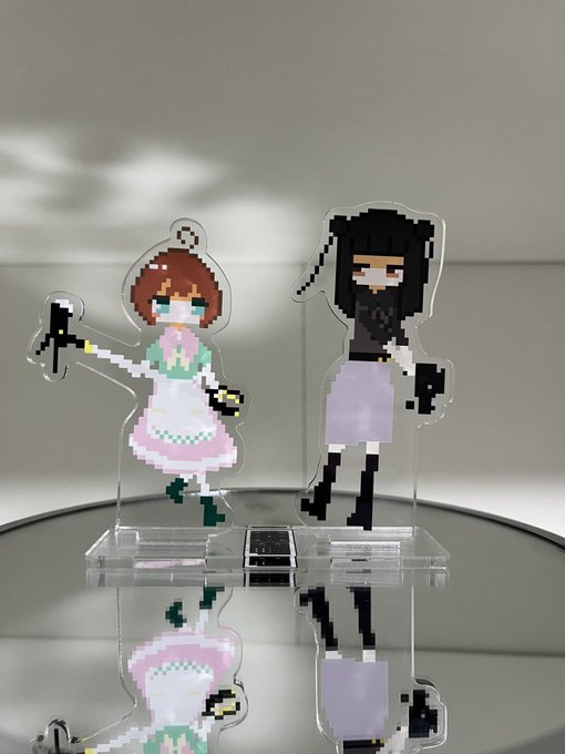 Two pixel art acrylic figures of anime-style girls with weapons stand on a reflective circular mirror, playfully illustrating the pairing of NANO and INOI cryptocurrencies as cute combat-ready companions.