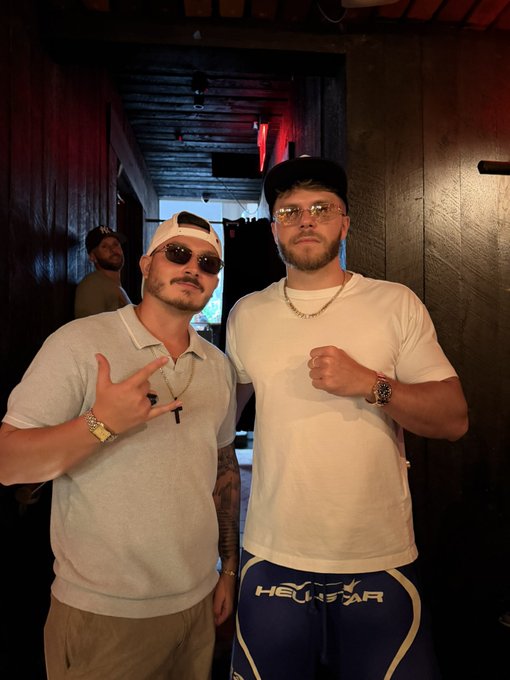 Two men posing for a photo in a dimly lit indoor setting; one wears sunglasses, a backwards cap, and a gray polo with a gold chain, while the other wears a white t-shirt, black cap, glasses, and Heydar-branded blue shorts.