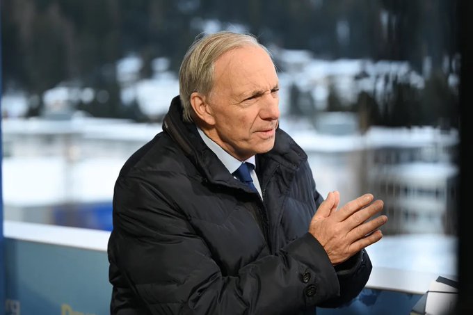 A man with light-colored hair, wearing a dark jacket over a white shirt and dark tie, stands outdoors rubbing his hands together, with a snowy landscape blurred in the background. The image accompanies an article about Ray Dalio's warning to reduce bond holdings and increase gold and Bitcoin allocations amid U.S. debt crisis concerns.