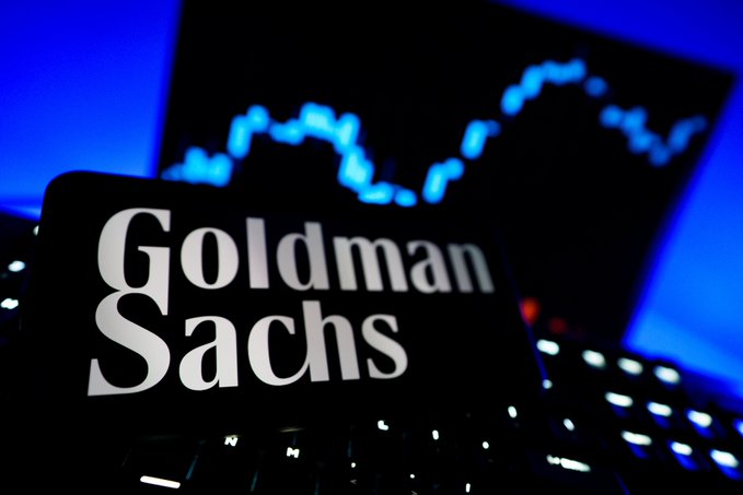 Goldman Sachs logo on a phone screen with a stock chart shown on a computer monitor in the background. The image relates to the post by visually referencing the prominent financial institution mentioned, Goldman Sachs, whose CEO is advocating for advancing the CLARITY Act.