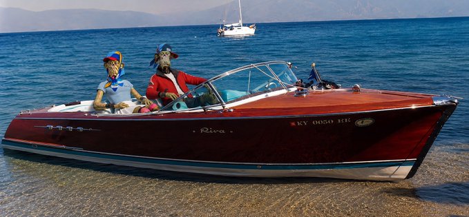 Two anthropomorphic goats ride a red wooden Riva boat on the ocean, with a sailboat visible in the background.