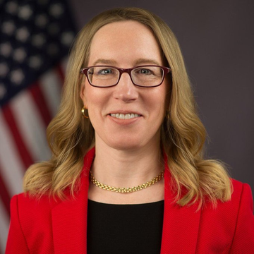 A professional headshot of a woman with shoulder-length blonde hair, glasses, a gold chain necklace, and a red blazer over a black top, posed in front of an American flag. The image accompanies the post about SEC Commissioner Hester Peirce's warning that crypto vaults and onchain lending strategies may be subject to federal securities laws, likely depicting Peirce herself delivering this regulatory message.
