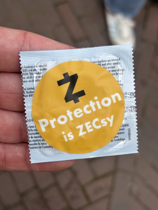 A hand holding a Zcash-branded condom with a yellow sticker reading 'Protection is ZECsy.'