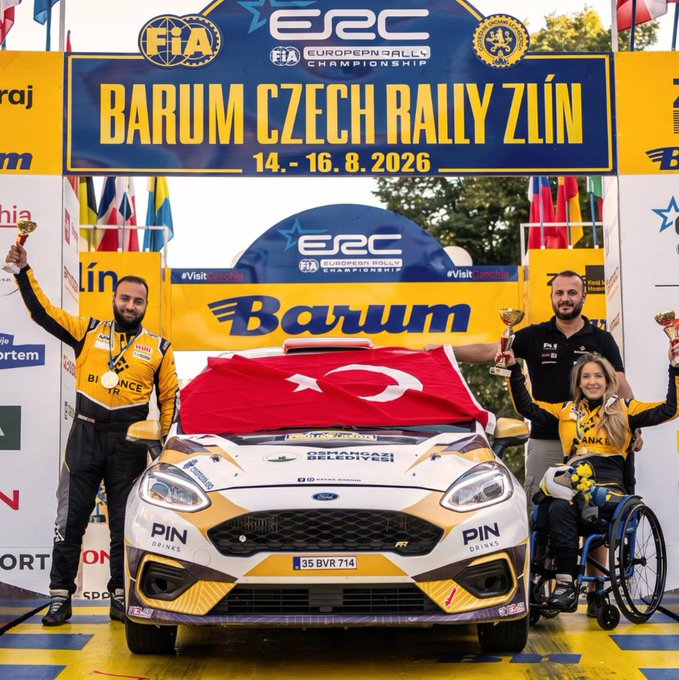 Kübra Denizci Keskin, a Team Binance TR athlete, celebrates on the podium alongside a co-driver holding trophies, with a Turkish flag draped over their gold and white Ford rally car (license plate "35 BVR 714") at the ERC Barum Czech Rally Zlín, dated 14-16.8.2026.