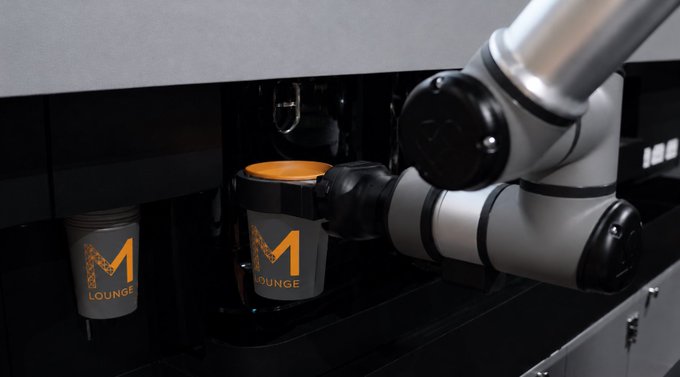 The image shows a coffee machine at a lounge that offers an exclusive experience for its shareholders. The coffee machine has two cups labeled "M" and "Lounge," indicating that this is a part of the lounge's unique offering, where customers can enjoy their favorite beverages in a futuristic setting. The coffee machine is connected to a robot arm, which is designed to serve drinks with precision and efficiency. This advanced technology ensures that guests receive their orders promptly and consistently, enhancing their overall experience at the lounge.