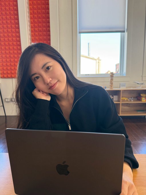 Shirley sits at a desk with an Apple MacBook, smiling at the camera with her chin resting on her hand. The setting appears to be a home or co-working space with red acoustic panels and a window in the background, suggesting a casual, focused work environment that aligns with her introduction as a founder building an agentic trading platform.