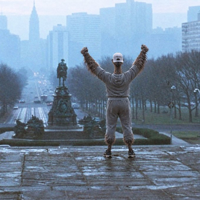 A person in a bird-like costume stands with arms raised on stone steps overlooking a city plaza featuring a prominent equestrian statue in foggy weather.