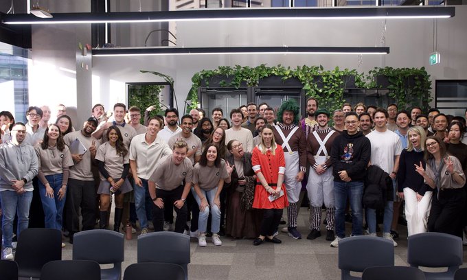 A group photo of approximately 40 people posing together in what appears to be an office or event space, with one person dressed in a costume featuring green hair and a plaid outfit.