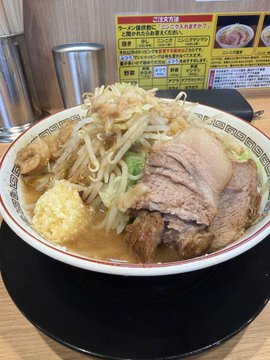 A large bowl of ramen topped with bean sprouts, sliced pork, green onions, and ginger, served on a wooden table with an informational sign in the background.
