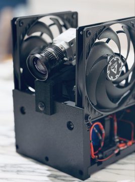 A close-up of a custom-built device featuring two large black computer fans mounted on a black metal frame, with a small camera lens visible between them and red and black wiring at the base.