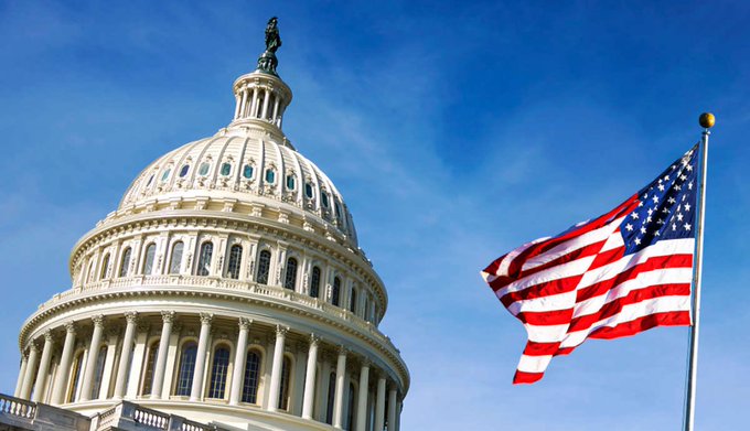 The U.S. Capitol building dome with a waving American flag set against a clear blue sky.