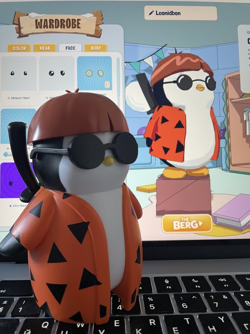 A figurine of a penguin character wearing sunglasses and an orange patterned jacket sits in front of a laptop displaying a wardrobe customization interface with options like "COLOR," "HEAD," "FACE," and "BODY."