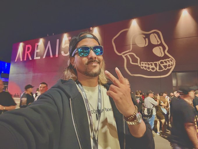 A man with long hair, blue reflective sunglasses, and a backwards cap takes a selfie while flashing a peace sign in front of an "AREA 15" wall featuring a large monkey skull graphic—visually fitting his "Apefest 2026" caption, which references the popular Bored Ape Yacht Club crypto community event.