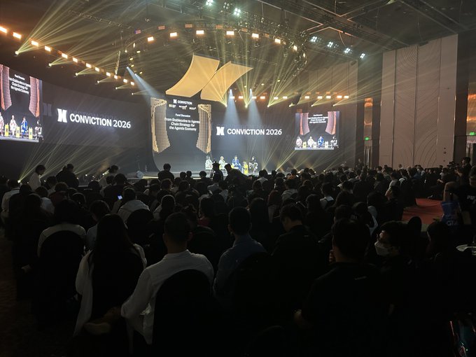 A large audience attends the 'CONVICTION 2026' conference in a darkened auditorium, with bright stage lights illuminating the stage and multiple screens displaying event branding and a speaker.
