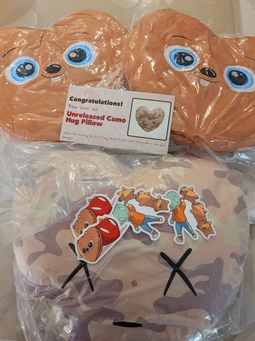 A collection of plush toys in plastic packaging, including two orange bear-like pillows with blue eyes and a camo-patterned pillow with cartoon bear stickers; a card reads 'Congratulations! You won an Unreleased Camo Hug Pillow'.