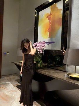 A woman in a long dark dress stands indoors near a console table decorated with flowers and artwork.